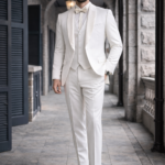 Man wearing an ivory three-piece suit with a white shirt and bow tie, walking confidently along a stone corridor.