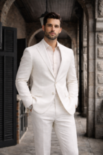 Male model wearing a white slim fit two-piece suit with open collar shirt, standing in a stone architectural outdoor setting.