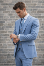 Man wearing a light blue three-piece suit adjusting his cuff against a brick wall background.