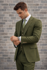 Man wearing an olive green three-piece suit adjusting his cuff against a brick wall background.