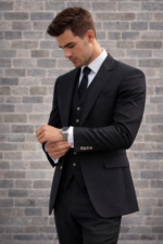 Man wearing a black three-piece suit adjusting his cuff against a brick wall background.
