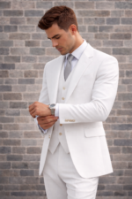 Man wearing a white three-piece suit adjusting his cuff against a brick wall background.